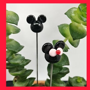 May include: Two black glass Mickey Mouse figurines with white eyes and red noses on metal stems. The figurines are in front of a green plant.