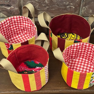Peut inclure: Quatre paniers ronds en tissu avec des poignées. Les paniers ont une base jaune avec des rayures rouges et un intérieur à carreaux rouges ou floral. Les poignées sont de couleur beige neutre. Les paniers sont sur une surface en bois.
