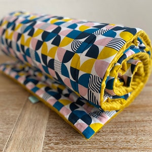 May include: A rolled up quilt with a geometric pattern in shades of pink, yellow, blue, and white. The quilt has a yellow border.