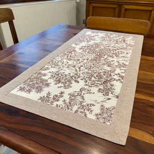 Peut inclure: Un chemin de table beige avec un motif floral brun et une bordure beige. Le chemin de table est sur une table en bois foncé.