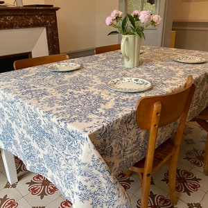 Op de afbeelding: Een wit tafelkleed met een blauw en wit bloemenpatroon, gelegd op een houten eettafel met vier stoelen. Het tafelkleed heeft een gegolfde rand en is versierd met een delicaat bloemenontwerp.