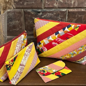 May include: A set of quilted pouches and a wallet. The designs feature diagonal stripes of red and yellow fabric, with floral, geometric, and gingham patterns. The pouches have red zippers. The largest pouch is approximately 30 cm wide.