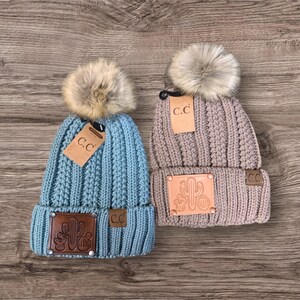 May include: Two knitted winter hats with faux fur pom poms. The hats are in blue and brown with a leather patch featuring a cactus design. The hats have a "C.C." logo on the side.