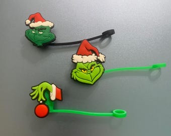 May include: Three Christmas-themed charms featuring the Grinch character. Two charms have the Grinch's face wearing a Santa hat, and one has the Grinch's hand holding an ornament. Each charm has a cord attached, with two black and one green.