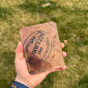 May include: A brown leather cardholder with the text "GENUINE HORWEEN 1905 SHELL CORDOVAN" in a circular stamp design. The cardholder has white stitching around the edges and is held in a hand.
