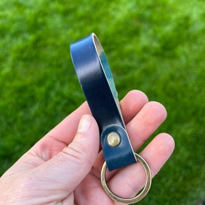 May include: A navy blue leather keychain with a brass-colored key ring and rivet. The leather strap is folded over and attached to the ring with a brass rivet. The background is green grass.