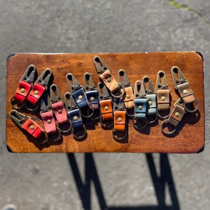 May include: A collection of leather keychains in various colors, including red, pink, blue, and brown. Each keychain features a metal clasp and a small leather loop with a metal rivet. The keychains are arranged on a wooden surface.