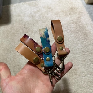 May include: Four leather keychains in various colors: tan, brown, burgundy, and blue with gold hardware. Each keychain has a snap closure and a metal clasp. The keychains are held in a hand.