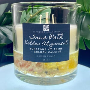 May include: A clear glass candle with a layered design. The candle features a white top layer, a middle layer with embedded stones, and a bottom layer with yellow glitter. The label reads "True Path Golden Alignment" with "Lemon Sugar" scent and is 16 oz.