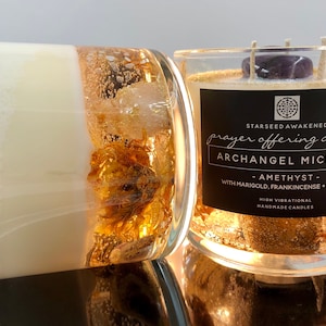 May include: Two glass candles with a layered design. The candles feature a white wax core, gold accents, and a decorative layer with gold flakes and dried flowers. One candle has a label that reads "Archangel Michael" and "Amethyst".