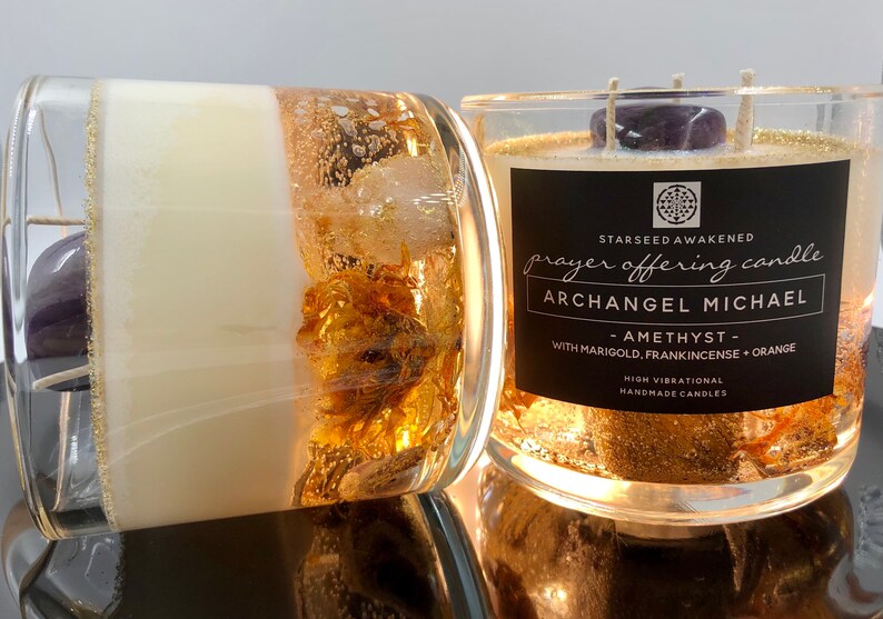 May include: Two glass candles with a layered design. The candles feature a white wax core, gold accents, and a decorative layer with gold flakes and dried flowers. One candle has a label that reads "Archangel Michael" and "Amethyst".