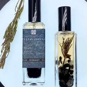 Könnte beinhalten: Zwei rechteckige Glasflaschen mit schwarzen Kappen. Eine Flasche hat ein Etikett mit der Aufschrift "Crystal Infusion", die andere enthält Kräuter und Kristalle. Die Flaschen stehen auf einer weißen Oberfläche mit einem Rosmarinzweig.