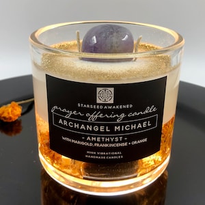 May include: A clear glass candle with gold glitter accents and a purple amethyst stone on top. The candle has a black label with white text that reads "prayer offering candle ARCHANGEL MICHAEL". The candle is handmade.