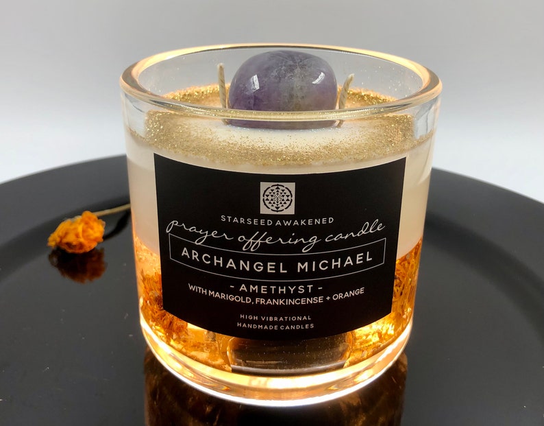 May include: A clear glass candle with gold glitter accents and a purple amethyst stone on top. The candle has a black label with white text that reads "prayer offering candle ARCHANGEL MICHAEL". The candle is handmade.