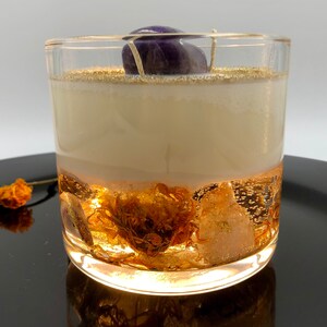 May include: A clear glass candle with a layered design. The bottom layer features gold-colored inclusions, while the middle layer is a creamy white. A purple stone sits atop the candle, and gold glitter accents the top.