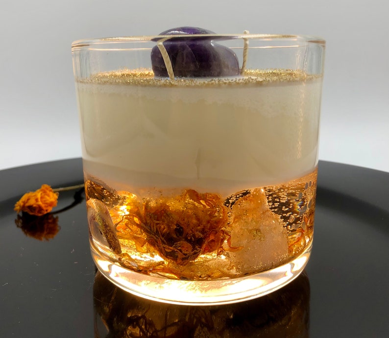 May include: A clear glass candle with a layered design. The bottom layer features gold-colored inclusions, while the middle layer is a creamy white. A purple stone sits atop the candle, and gold glitter accents the top.