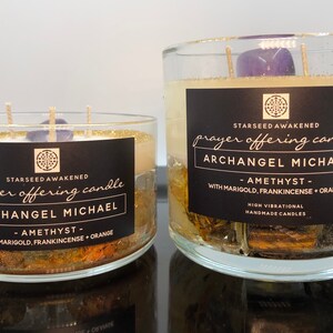 May include: Two glass prayer offering candles labeled "Archangel Michael" with amethyst, marigold, frankincense, and orange. The candles have a black label with white text and gold glitter around the top.