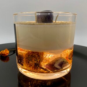 May include: A clear glass candle with a layered design. The bottom layer features dried flowers and crystals, followed by a creamy wax layer. A purple crystal sits atop the candle. The candle is on a black surface.