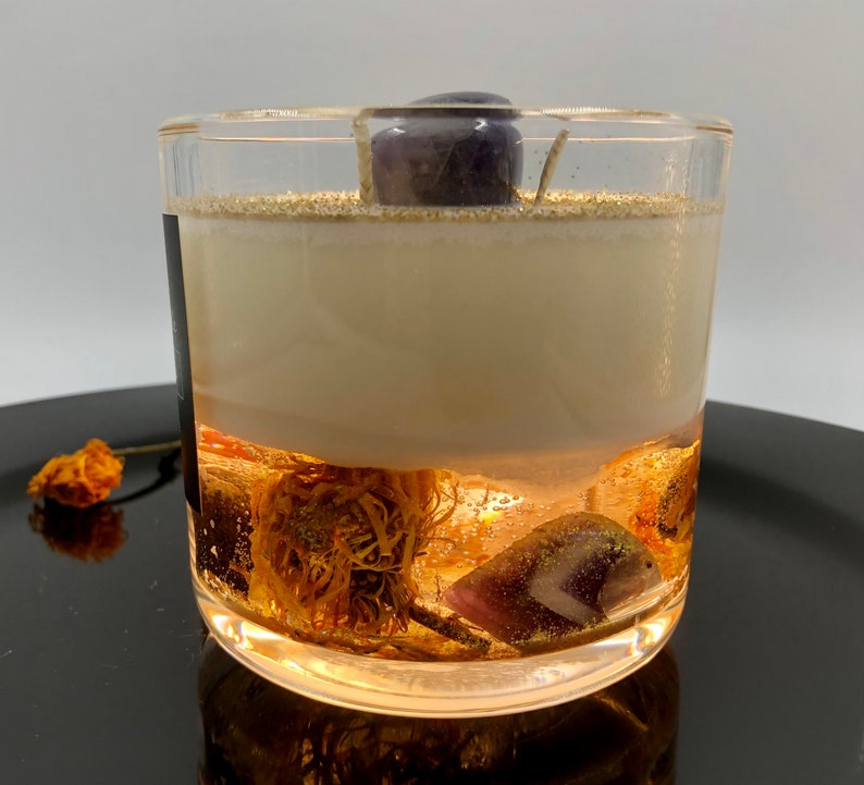 May include: A clear glass candle with a layered design. The bottom layer features dried flowers and crystals, followed by a creamy wax layer. A purple crystal sits atop the candle. The candle is on a black surface.
