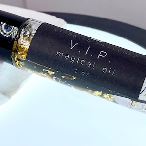 以下が含まれることがあります： 金色のフレークと液体が入った、黒いキャップが付いた透明なガラス製のロールオンボトル。ボトルには「V.I.P. magical oil 1 oz.」と書かれた黒いラベルが付いています。ボトルは白い表面に置かれています。