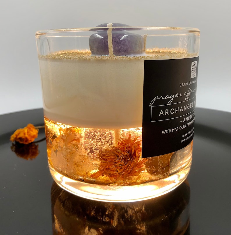 May include: A clear glass candle with a purple amethyst stone on top. The candle has a white wax layer and a clear gel layer with gold flakes and dried flowers. The label reads "prayer offering" and "Archangel".
