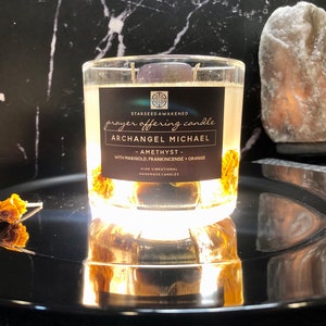May include: A clear glass candle labeled "Archangel Michael" with the text "prayer offering candle". The candle contains a purple stone, marigold, and orange. The candle sits on a reflective black surface.