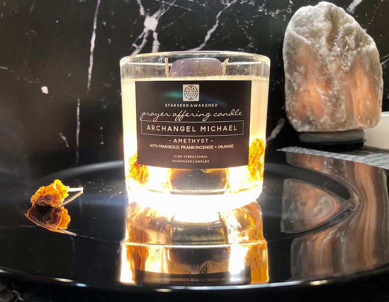 May include: A clear glass candle labeled "Archangel Michael" with the text "prayer offering candle". The candle contains a purple stone, marigold, and orange. The candle sits on a reflective black surface.