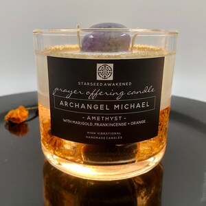 May include: A clear glass candle with a black label that reads "Prayer Offering Candle" and "Archangel Michael." The candle contains a purple amethyst stone and gold flakes. The text on the label also says "Amethyst - With Marigold, Frankincense + Orange."