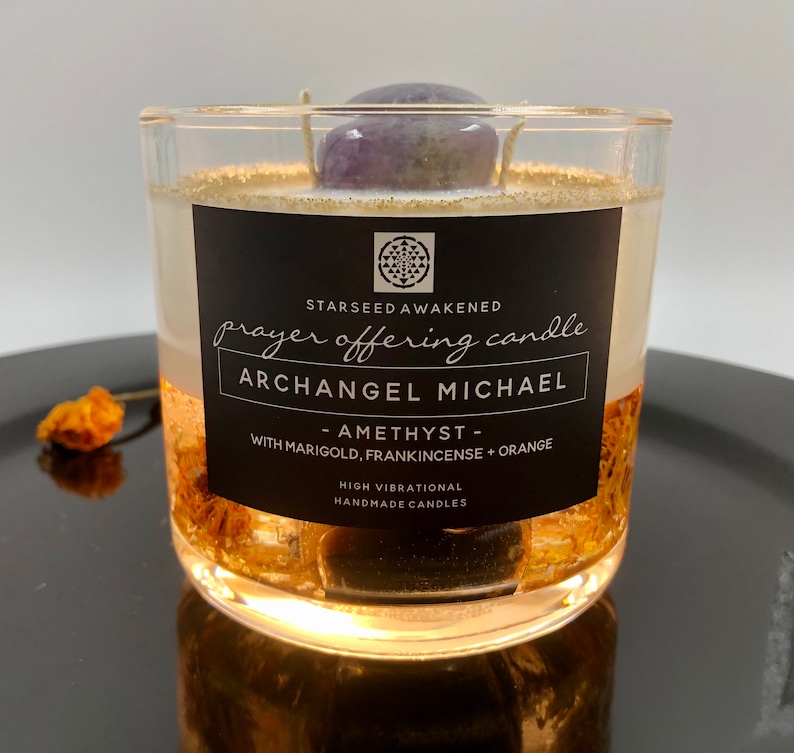 May include: A clear glass candle with a black label that reads "Prayer Offering Candle" and "Archangel Michael." The candle contains a purple amethyst stone and gold flakes. The text on the label also says "Amethyst - With Marigold, Frankincense + Orange."