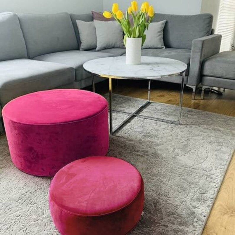 May include: Two round pink velvet ottomans, one larger than the other, are placed on a gray rug in front of a gray sectional sofa. A marble-topped coffee table with a chrome frame sits between the ottomans and the sofa. A bouquet of yellow tulips in a white vase sits on the coffee table.