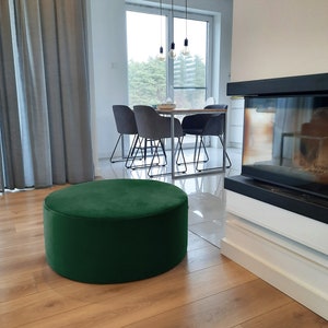 May include: A round, emerald green velvet ottoman sits on a light brown hardwood floor. The ottoman is the focal point of a modern living room, with a dining table and chairs visible in the background. A fireplace and window are also in the background.