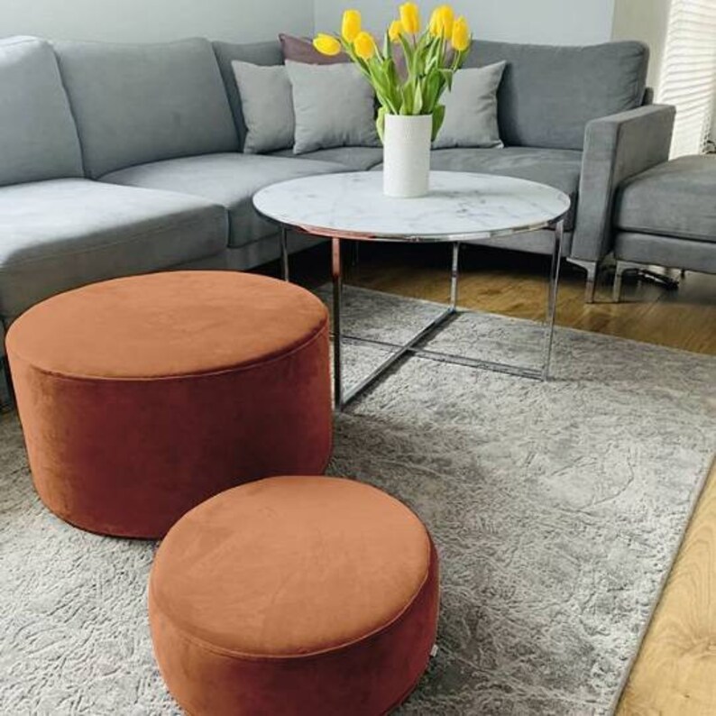 May include: A modern living room setup featuring a gray sectional sofa, a round marble-top coffee table with a chrome base, and two rust-colored velvet ottomans. A vase of yellow tulips adds a pop of color. The room is decorated with a patterned gray rug.