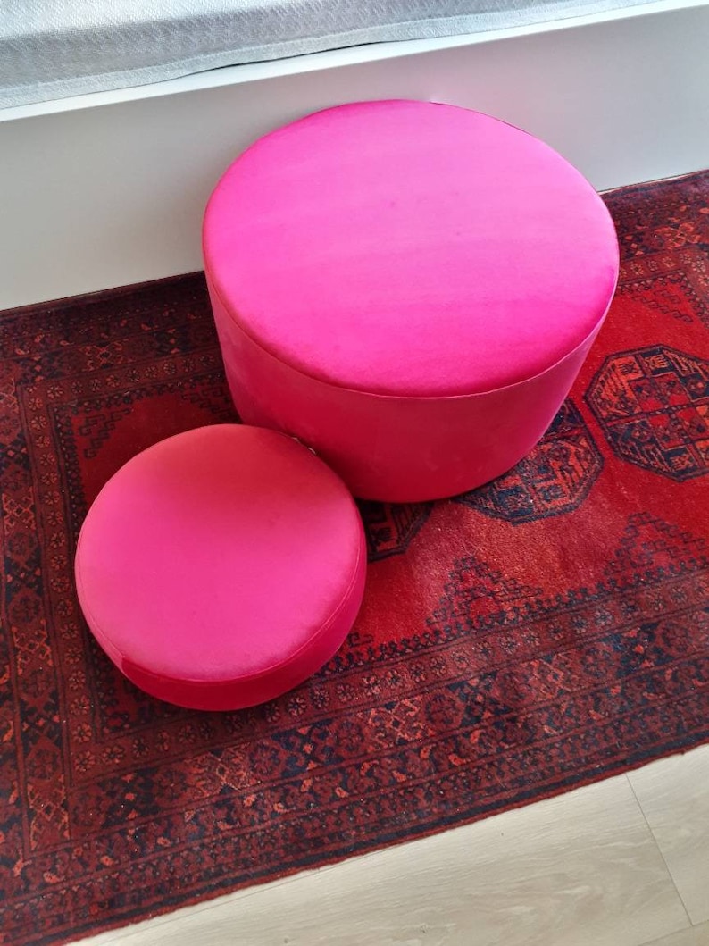 May include: Two round pink velvet ottomans on a red patterned rug. The larger ottoman is in the background and the smaller ottoman is in the foreground.