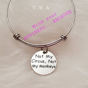 May include: A silver-toned bangle bracelet with a round charm. The charm is engraved with the words "Not My Circus, Not My Monkeys." The bracelet also has the words "Mind your BUSINESS...BECAUSE" in pink text.