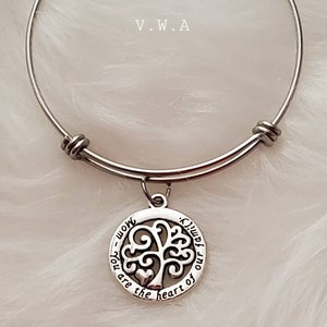 May include: A silver-toned bangle bracelet with a circular charm featuring a tree design. The charm has the words "Mom - you are the heart of our family" engraved around the tree. The bracelet is set against a soft, white background.
