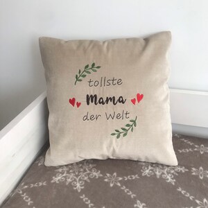 May include: Beige pillow with green leaves and red hearts embroidered on it. The text "tolle Mama der Welt" is embroidered on the pillow.