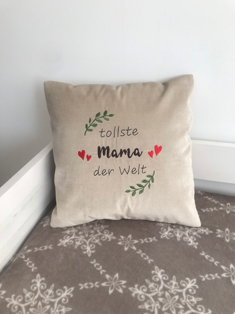 May include: Beige pillow with green leaves and red hearts embroidered on it. The text "tolle Mama der Welt" is embroidered on the pillow.
