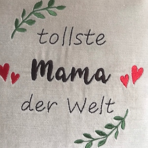 May include: A white cushion with green embroidered leaves and red hearts. The cushion has black embroidered text that reads "tollste mama der Welt".