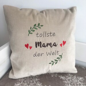 May include: Beige pillow with embroidered text in brown and red that reads "tolle Mama der Welt" with green leafy sprigs on either side.