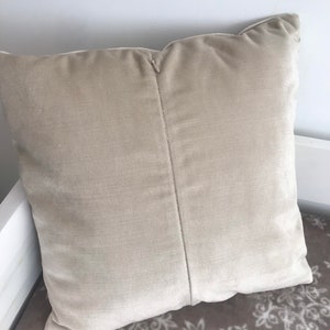 May include: A beige velvet cushion with a seam down the centre. The cushion is lying on a brown and white patterned surface.