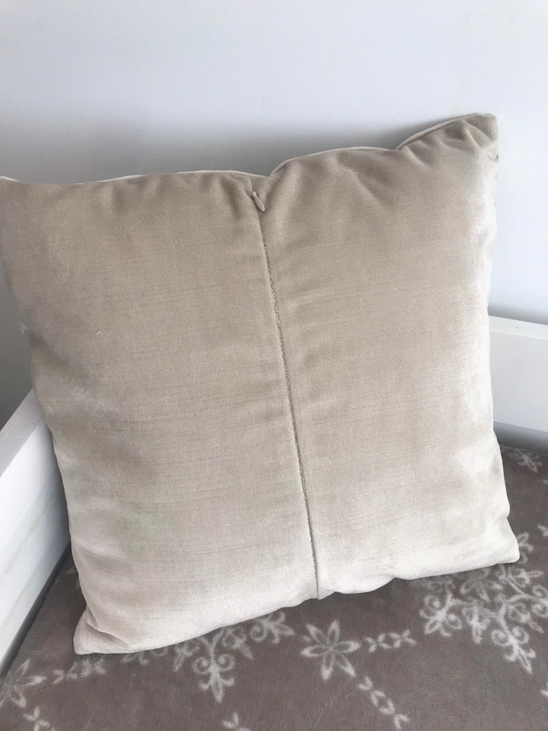 May include: A beige velvet cushion with a seam down the centre. The cushion is lying on a brown and white patterned surface.