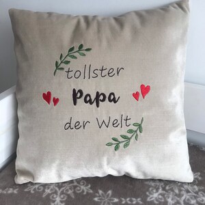 May include: Beige throw pillow with embroidered text "tollster Papa der Welt" in black with two red hearts and green leafy branches.