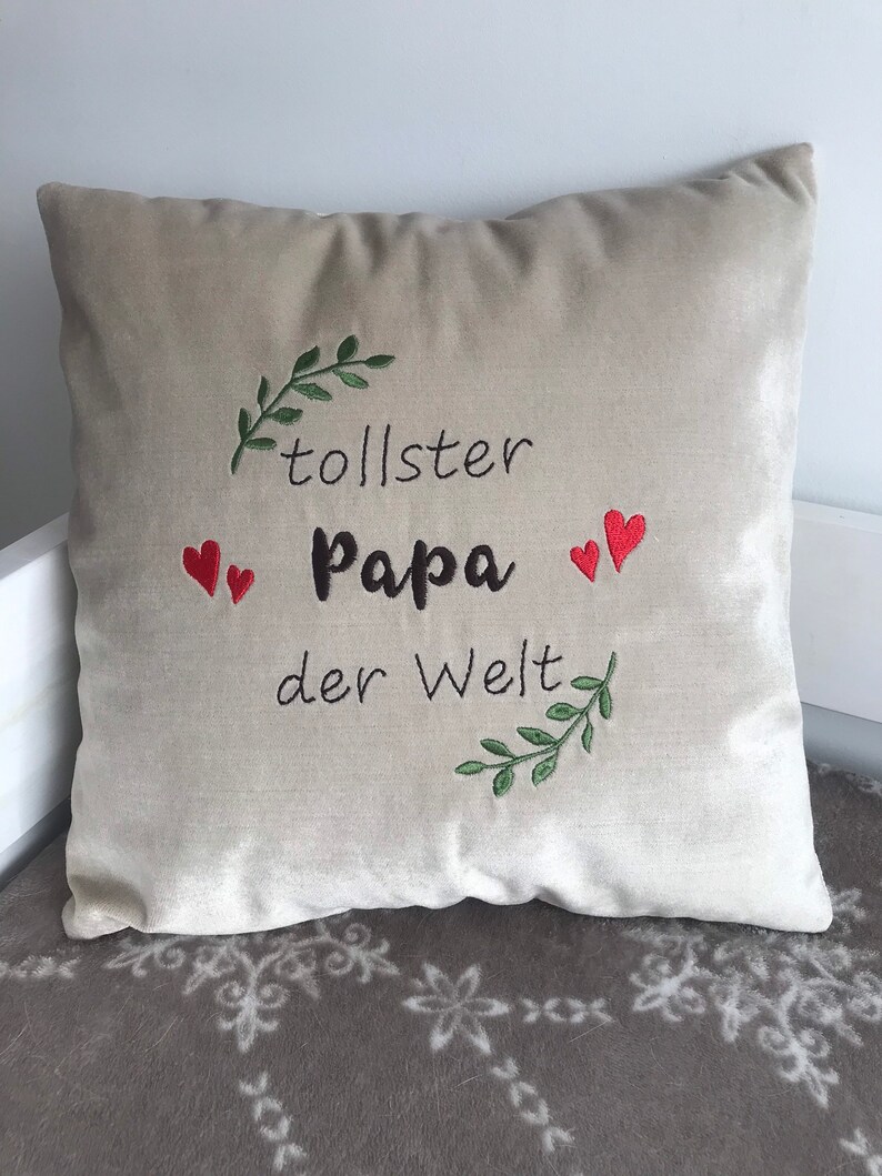 May include: Beige throw pillow with embroidered text "tollster Papa der Welt" in black with two red hearts and green leafy branches.