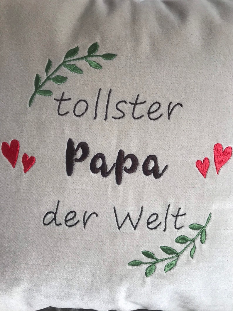 May include: A white pillow with green embroidered leaves and red hearts around the text "tollster Papa der Welt".