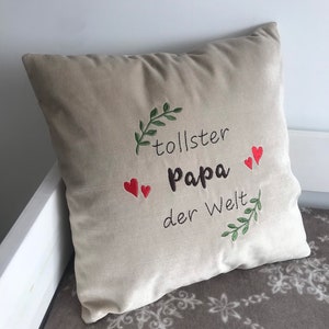 May include: Beige pillow with green leaves and red hearts embroidered on it. The text "tollster Papa der Welt" is embroidered on the pillow.