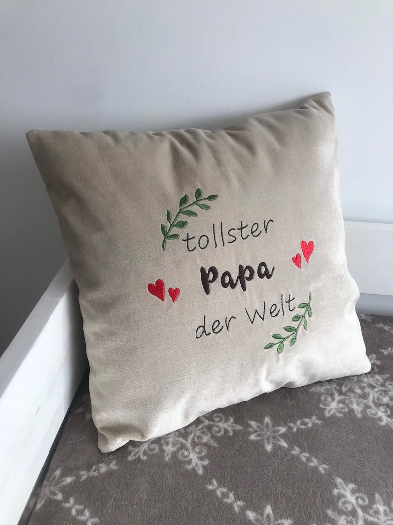 May include: Beige pillow with green leaves and red hearts embroidered on it. The text "tollster Papa der Welt" is embroidered on the pillow.