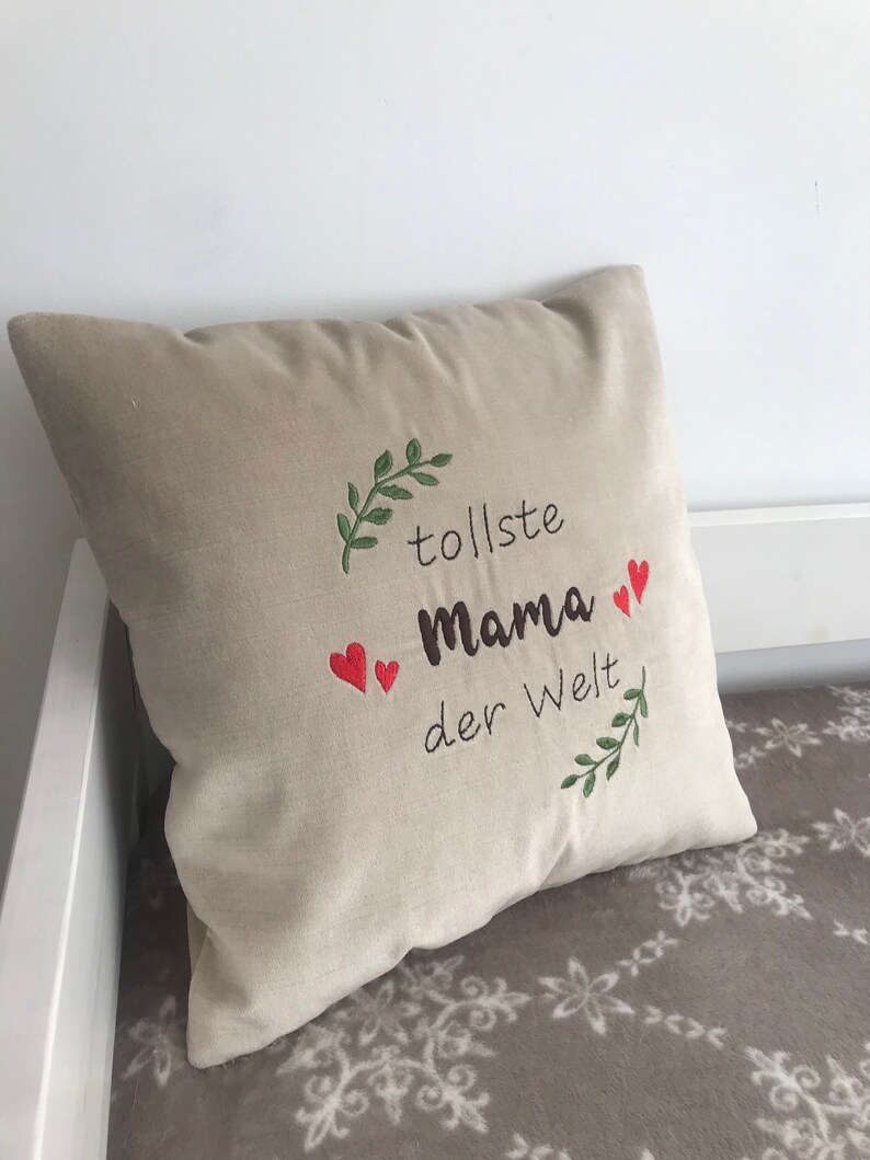 May include: Beige pillow with embroidered green leaves and red hearts. The text "tollste Mama der Welt" is embroidered on the pillow.