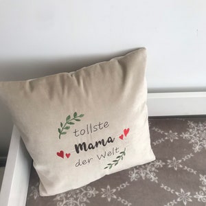 May include: Beige throw pillow with embroidered green leaves, red hearts, and the text "tollste Mama der Welt".