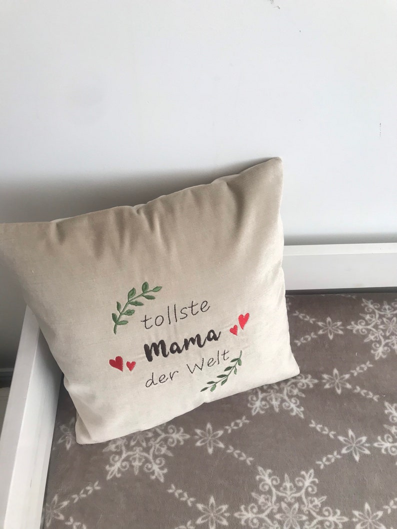 May include: Beige throw pillow with embroidered green leaves, red hearts, and the text "tollste Mama der Welt".