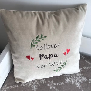 May include: Beige throw pillow with embroidered green leaves and red hearts. The text "tollster Papa der Welt" is embroidered in brown on the pillow.